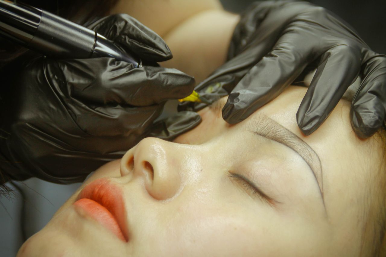 home-hero-1 A detailed view of eyebrow microblading procedure with gloved hands and closed eyes.